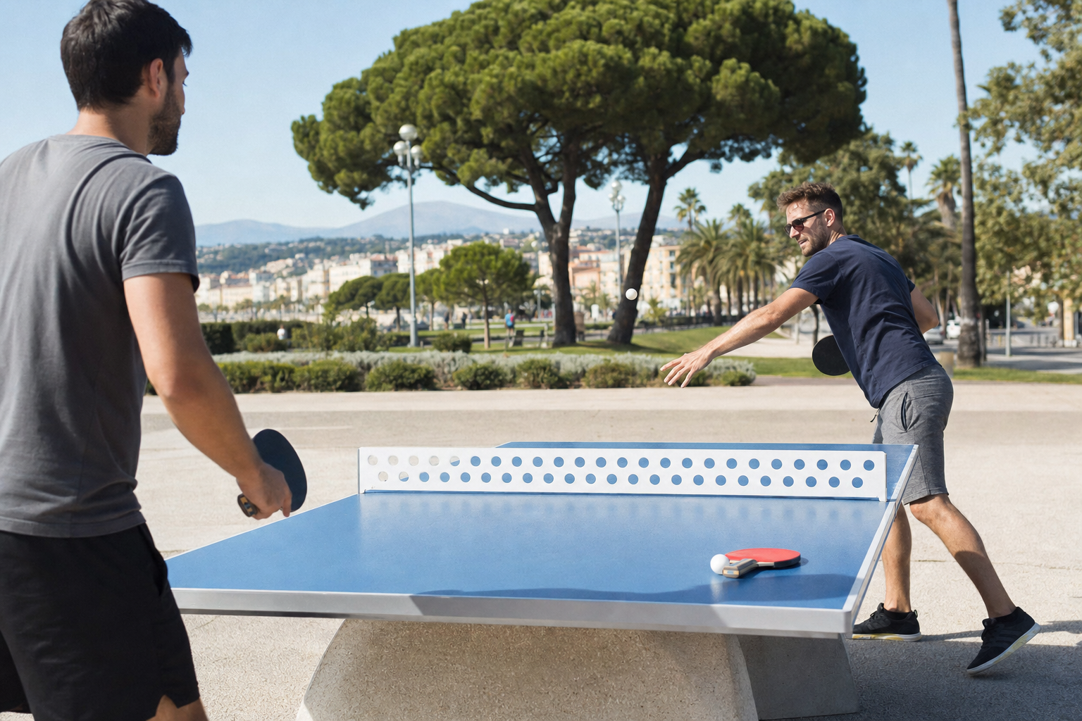 Table de ping-pong publique bleue dans un parc méditerranéen de Nice, avec deux adultes jouant en plein soleil.