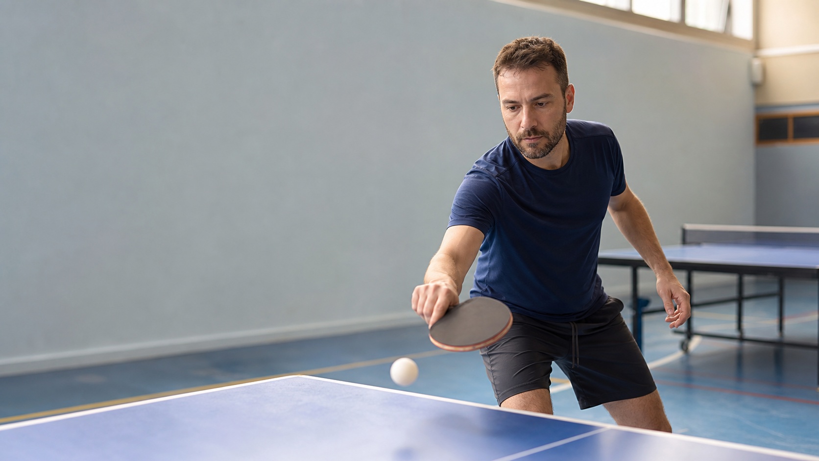 Débutant concentré réalisant un coup droit à une table de tennis de table dans une salle de sport lumineuse.