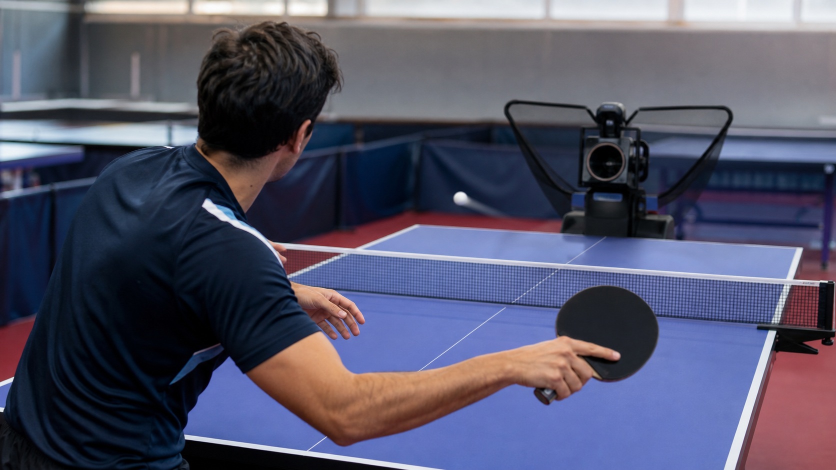 Joueur de tennis de table s’entraînant face à un robot lance-balles sur une table bleue dans une salle de sport.