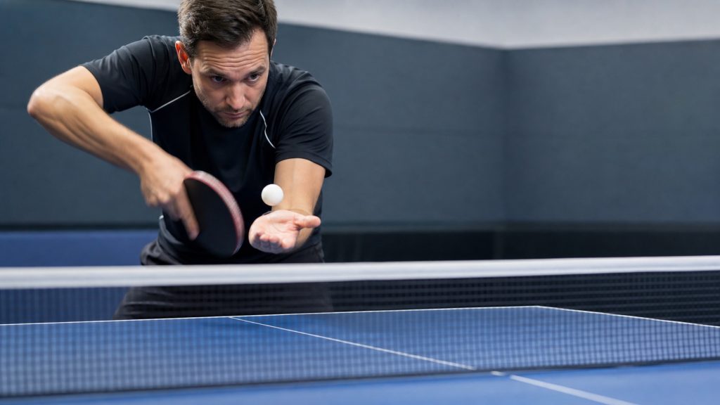 Joueur de tennis de table réalisant un service coupé avec effet, balle blanche au-dessus de la main et raquette en mouvement devant une table bleue.