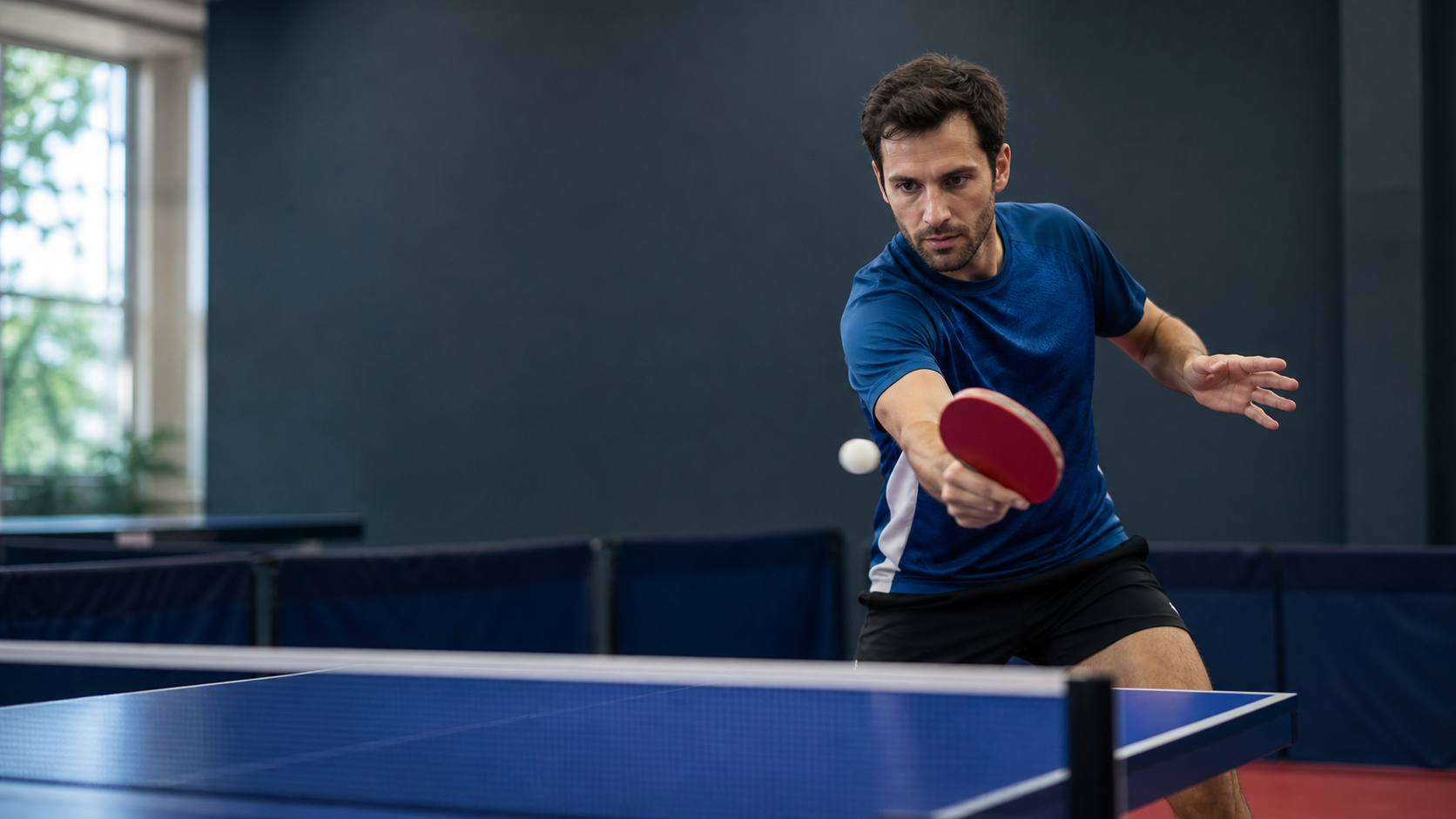 Joueur de tennis de table réalisant un revers en topspin sur une table bleue dans une salle de sport, avec espace dégagé à gauche.